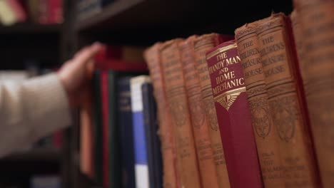 La-Mano-Recorre-Libros-Clásicos-En-La-Biblioteca,-Eligiendo-Agarrar-Un-Libro-Con-Tapa-Roja