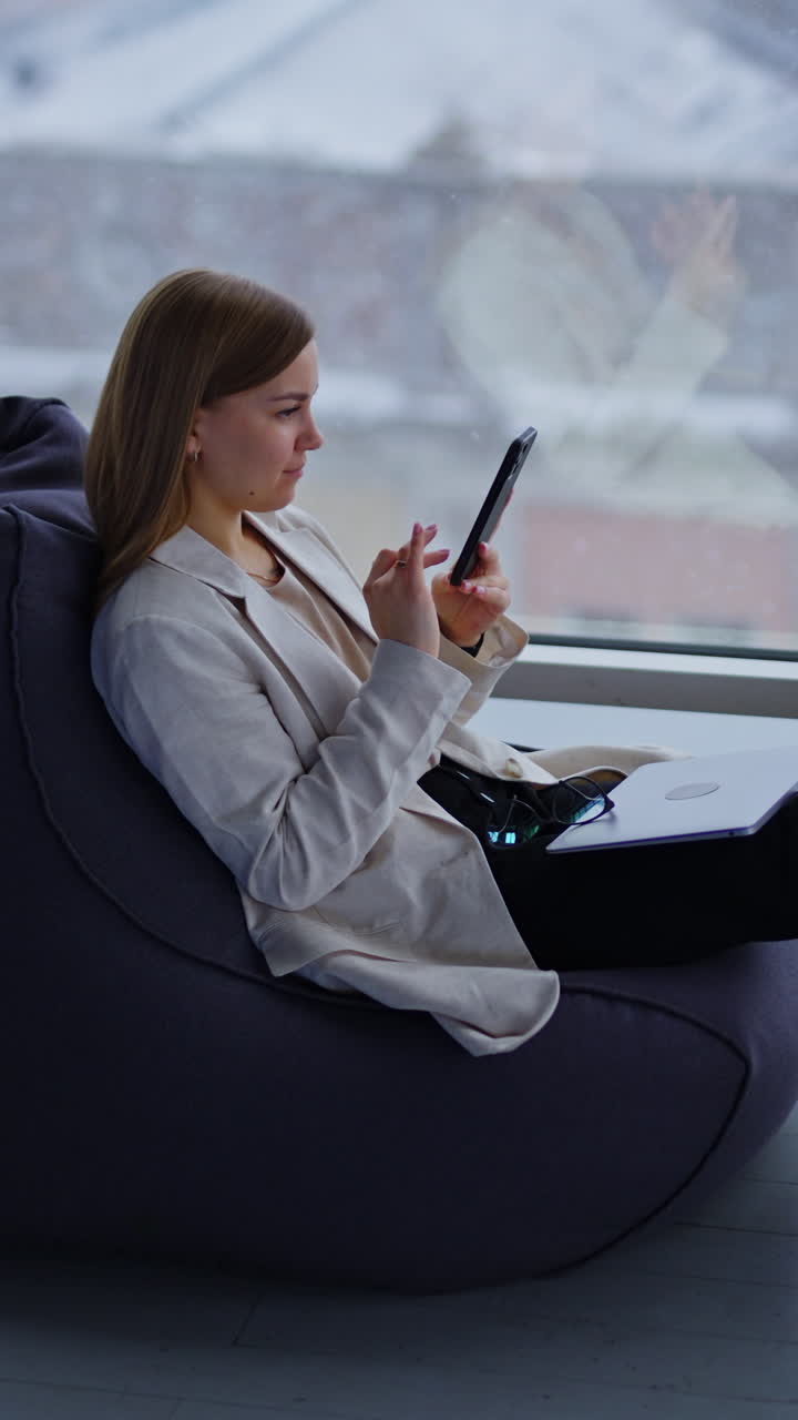 Calm relaxed lady looks at her phone sitting at chair stretching legs in front of her. Woman has laptop and glasses on her laps. Snow falling behind the window at backdrop. Vertical video