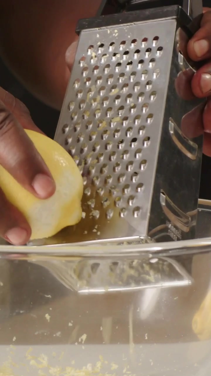 Video vertical: Rallando la cáscara de un limón, con manos inclinando un rallador sobre un cuenco en la cocina, recogiendo la ralladura para un plato.