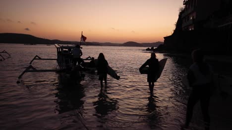 silhouette of surfers with surfboard leaving gerupuk beach by traditional boat at sunset in lombok, indonesia. - wide, slow motion