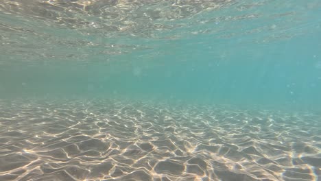 clear underwater footage shows rippled sand illuminated by sunlight, with gentle water movement and a tranquil, blue aquatic environment. wide, steady camera shot