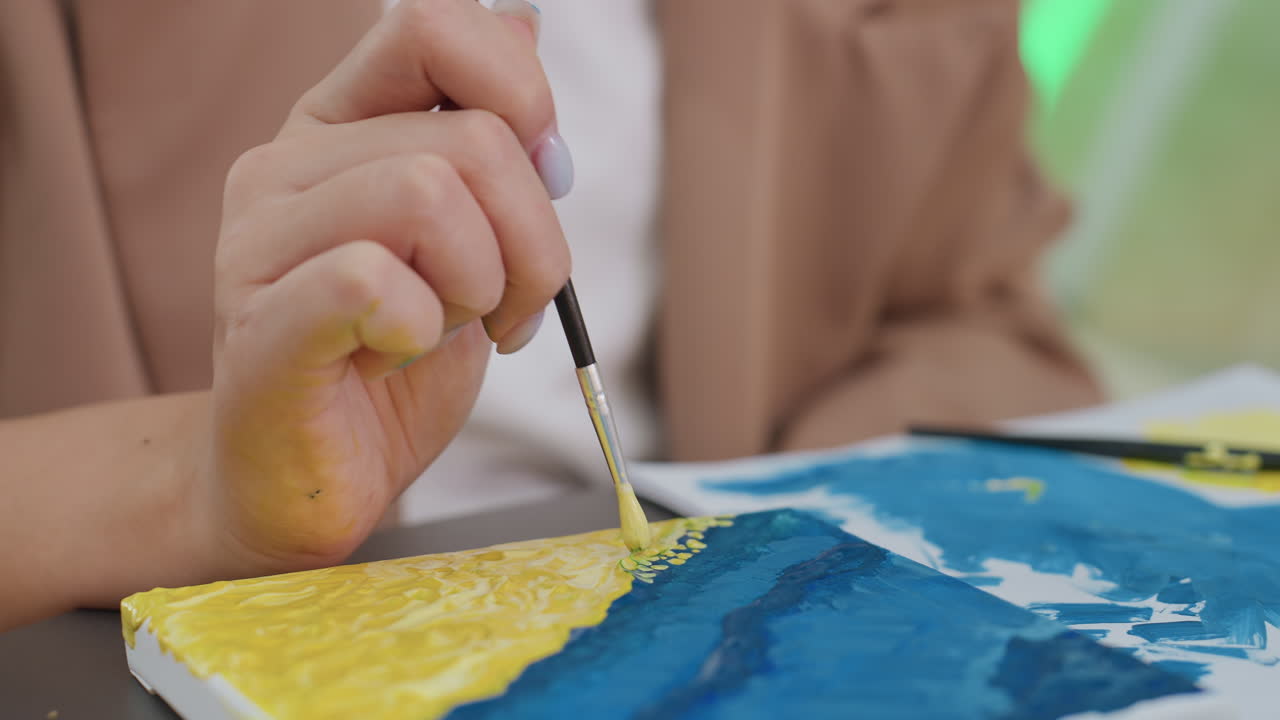 Primer plano de unas manos blancas femeninas aplicando reflejos blancos a lo largo del borde de una ola sobre un lienzo azul y amarillo, pinceladas precisas, con la manga de una chaqueta pulcra visible, luces suaves de estudio, ambiente de instrucción creativa enfocada