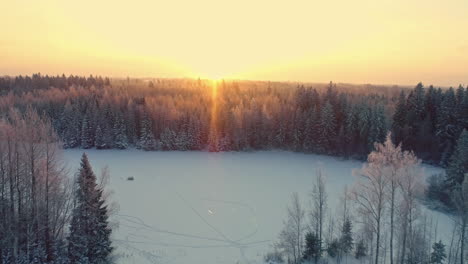 Aerial-drone-forward-moving-shot-over-a-frozen-lake-beside-rectangular-cottage-and-barrel-sauna-on-a-snow-covered-winter-landscape-at-sunrise