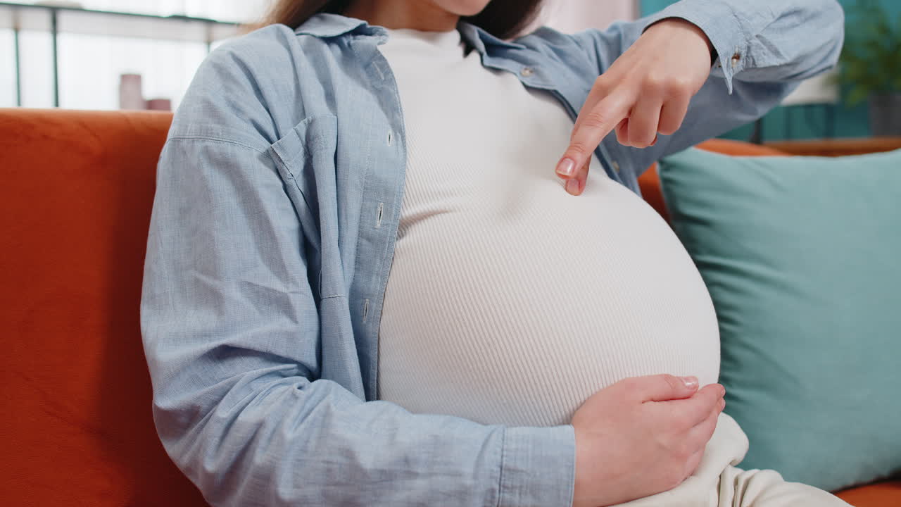임신 중인 젊은 여성은 태어나지 않은 아이와 함께 놀고 있는 아기를 기다리고 있는 그녀의 큰 배 위에 손가락을 고 있습니다.