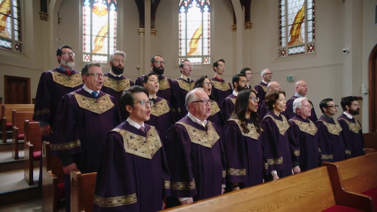 Inizio di una canzone, coro che apre le bocche e canta in un santuario con vetrate colorate, in abiti viola.
