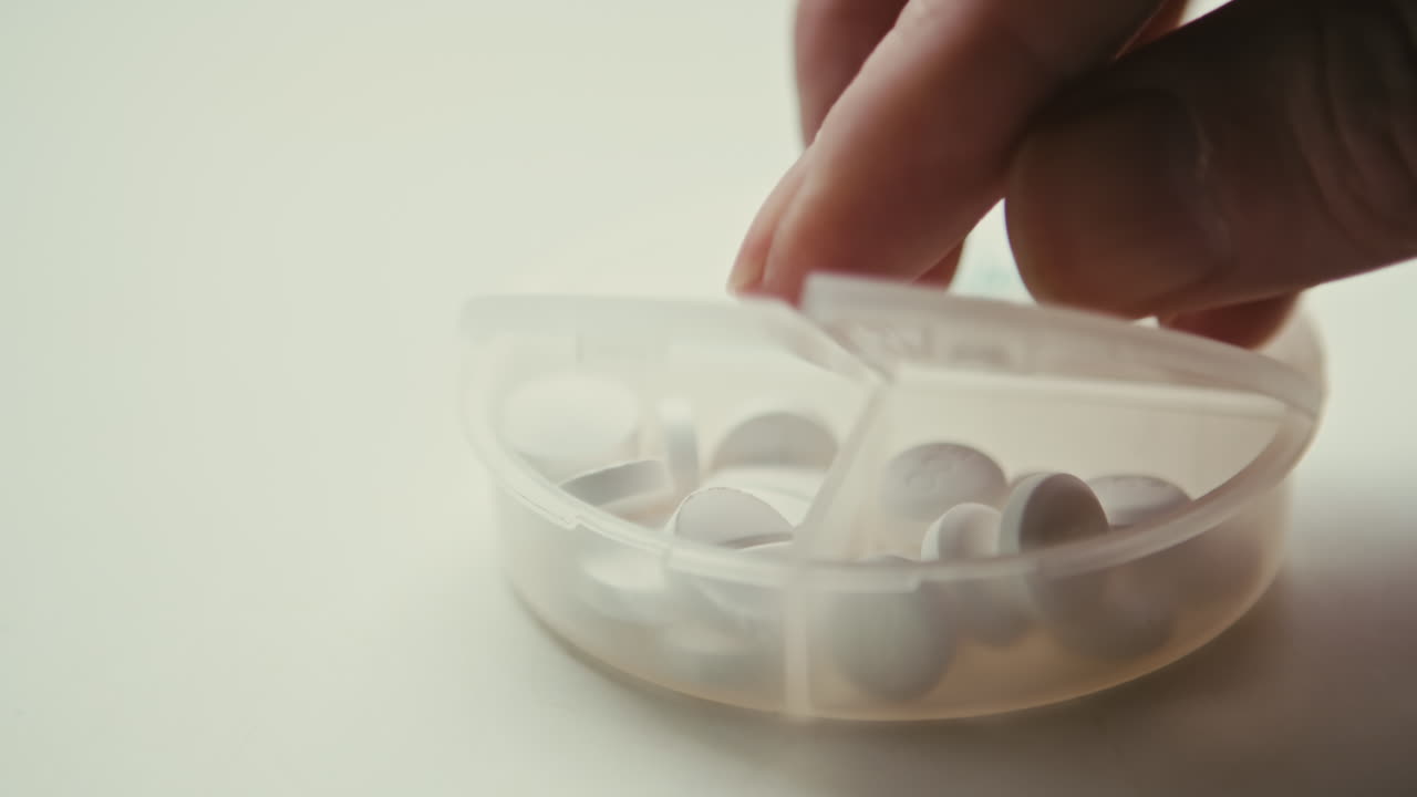 Close up of a person opening a clear plastic pill box to retrieve white tablets. Concept for daily routine, supplement intake, or chronic care