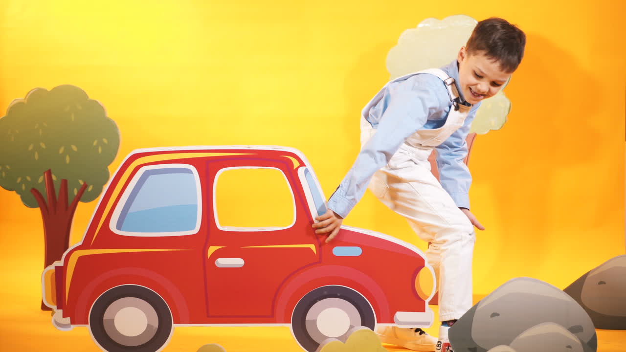 Happy kid in the children's studio with toyful decor on the yellow background. Funny little boy comes to the studio, sits on carton car and raises it up by his hands.