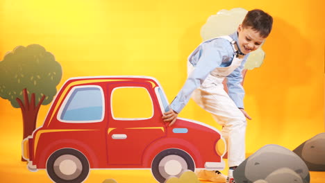 happy kid in the children's studio with toyful decor on the yellow background. funny little boy comes to the studio, sits on carton car and raises it up by his hands.