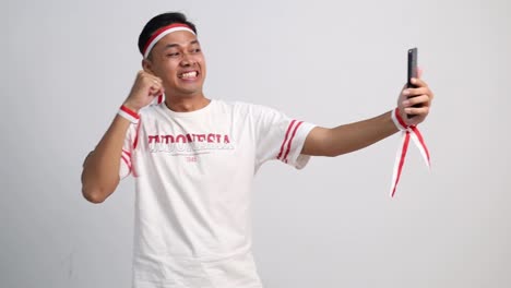 smiling young asian man taking selfie on mobile phone with make peace sign. indonesian independence day celebration concept
