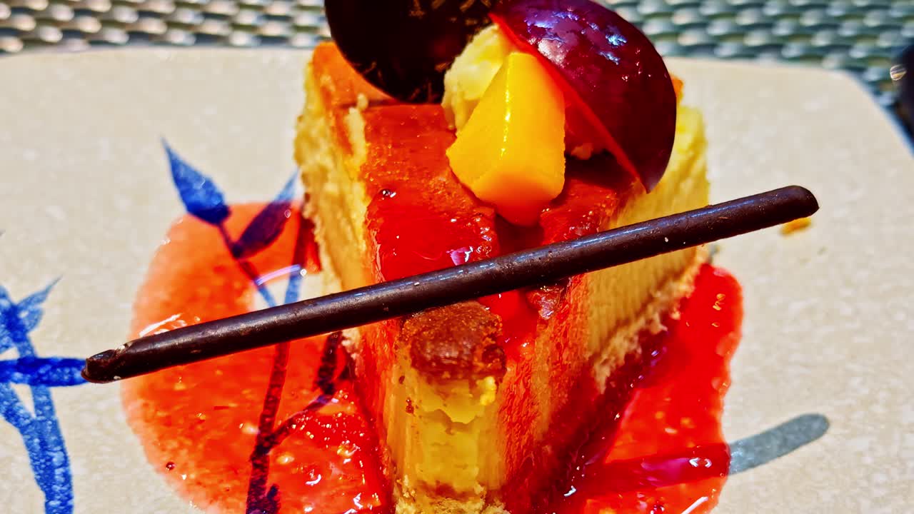 Close-up of creamy cheesecake slice topped with fruit, red glaze, and chocolate stick on a dessert plate