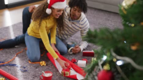 Video-of-happy-diverse-couple-in-christmas-antlers-and-santa-hat-wrapping-gifts-at-home