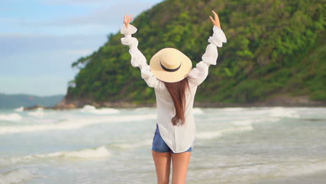 a young woman with her back to the camera as she walks through the incoming surf spontaneously throws her arms in the air in joy