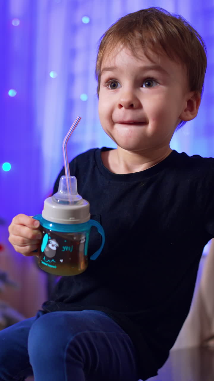 Small toddler boy with a cup in hand sits on the stairs. Cute kid in the room with Christmas tree. Vertical video