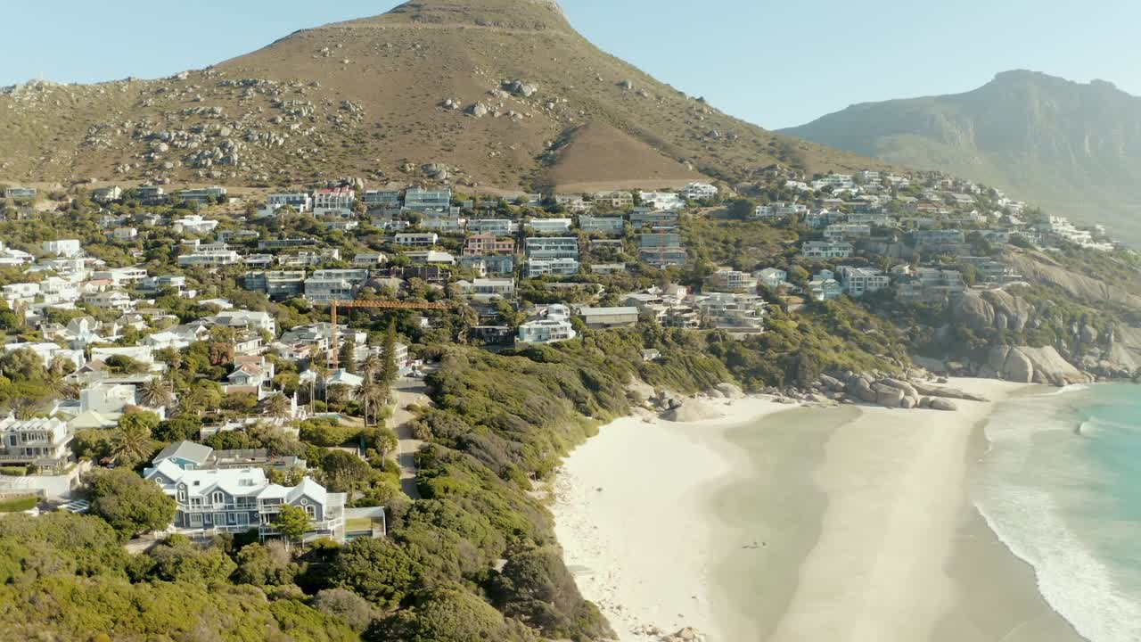 Drone view captures crowded town nestled between majestic, arid mountain ranges and scenic beach in Iceland. The rugged mountains contrast with town's dense architecture