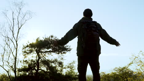 silhouette of man with backpack in wilderness pulling up arms in front of sunlight in nature