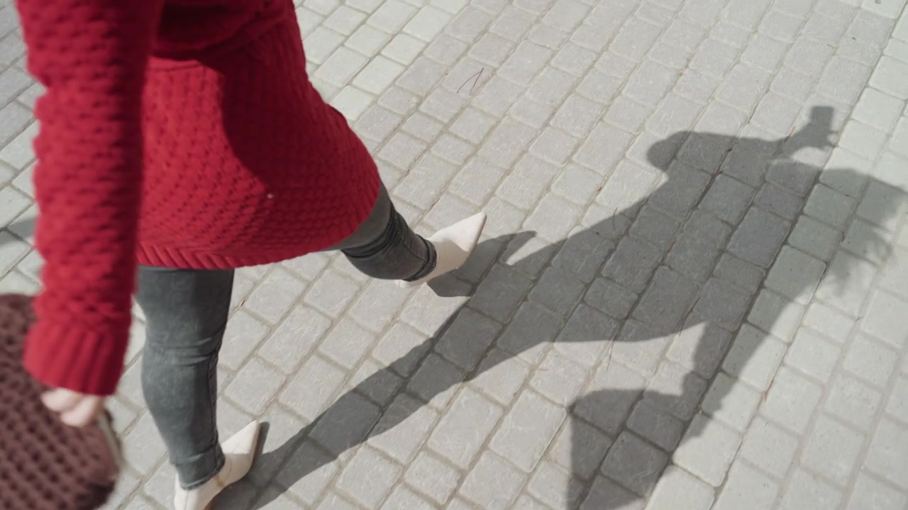 Close up of woman wearing red sweater and gray pants walking on light colored brick pavement holding brown knitted item. Her shadow is visible alongside her as woman moves through outdoor