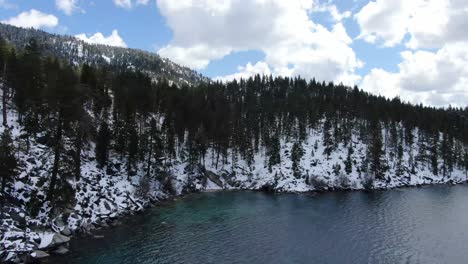 a slow revealing 4k drone cinematic shot of the shoreline of lake tahoe on a sunny day with a dark coast that is filled with pine trees as you see the sierra nevada mountains of nevada and california.