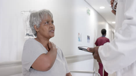 un médico masculino serio y diverso con una tableta hablando con un paciente mayor en la cama en cámara lenta, inalterado