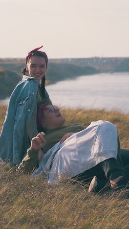 pretty woman with girlfriend spends time on autumn grass near camp against wide tranquil river and evening sky