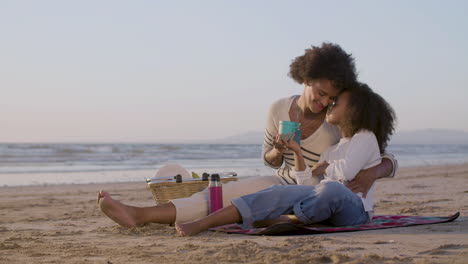 amorosa madre e hija bebiendo té y hablando mientras se sientan en una manta y hacen un picnic en la playa