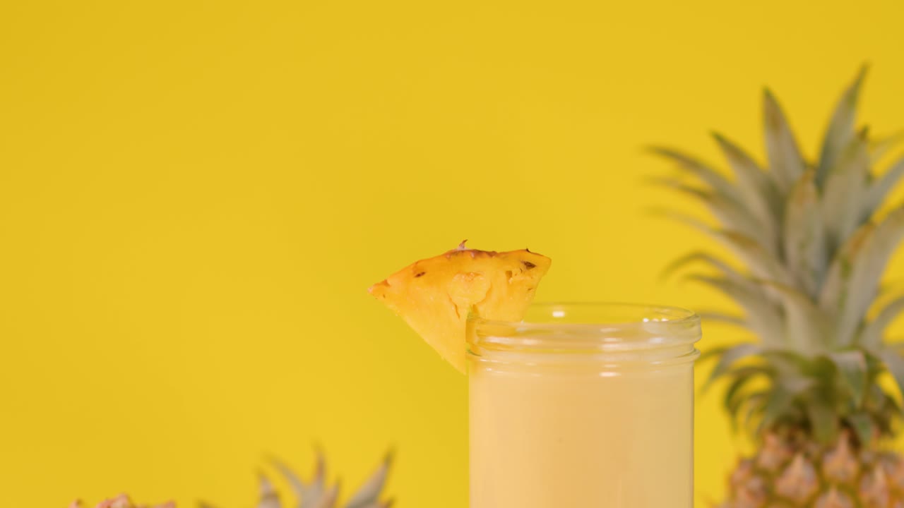 Hand adds pineapple-shaped stirrer to creamy smoothie in glass, bright yellow backdrop, tropical atmosphere