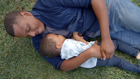 mixed race dad lies tickling young son on the grass in park