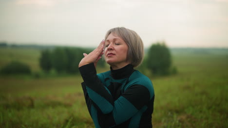 woman in green and black suit practicing cow face yoga pose outdoors in a misty grassy field, hands raised in a meditative gesture, with a soft blurred background of nature and trees