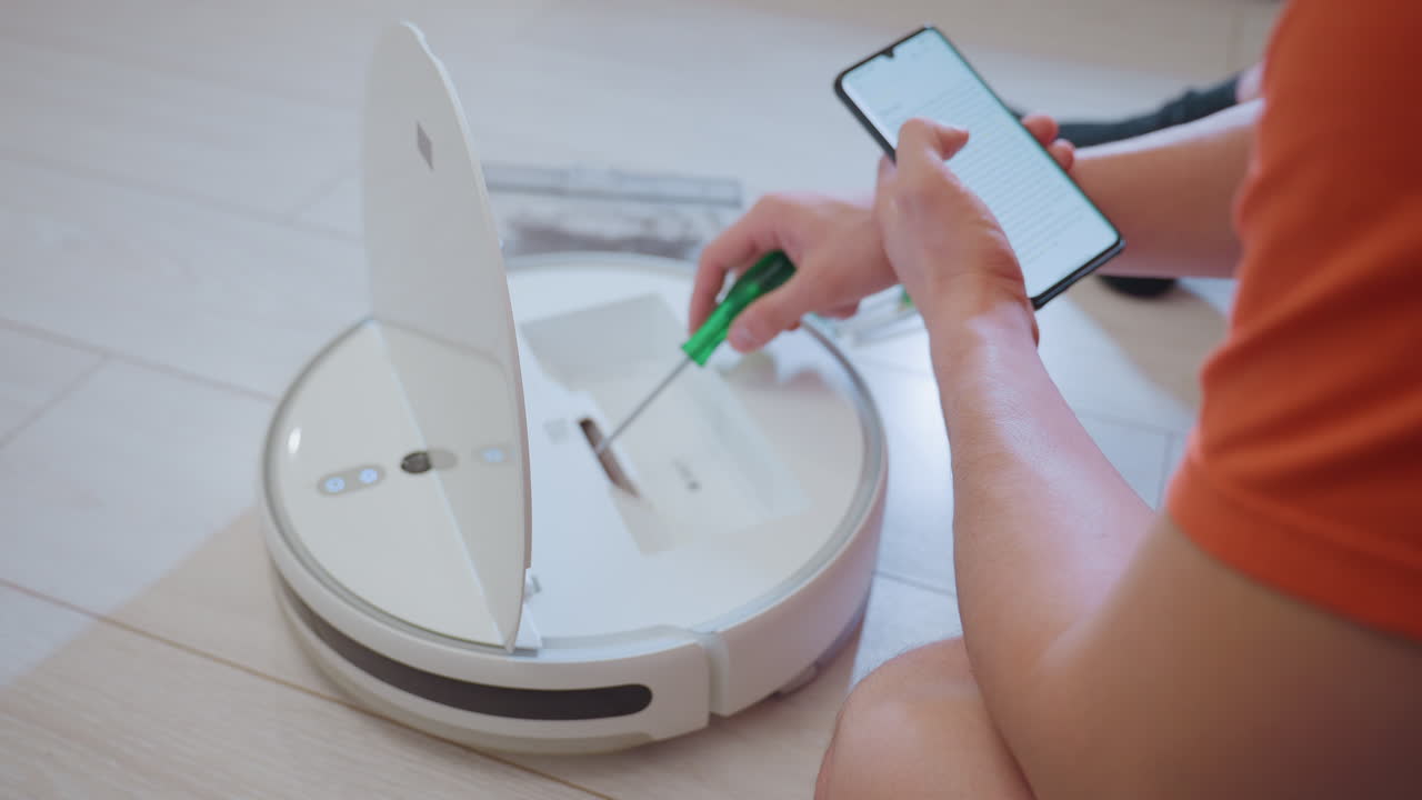 Close up of engineer seated on floor using phone for guidance while repairing opened robot vacuum with tools beside, focusing on fixing device, improving household cleaning and maintenance efficiency