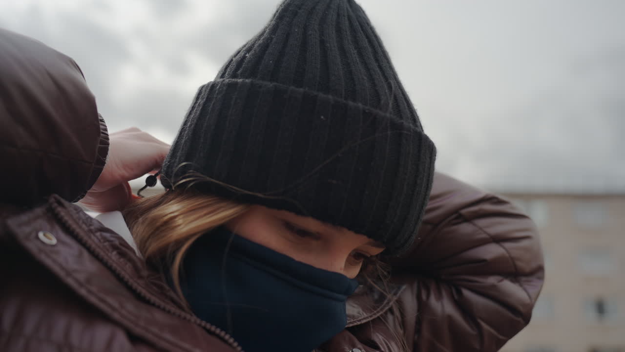 바람이 부는 회색 하늘 아래 옥상에서 비니와 스카프를 매고 있는 백인 여성, 레이어드 코트, 클로즈업된 손으로 머리카락을 정리하고 니트 텍스처, 차분하고 보호적인 분위기, 배경의 동네 건물들