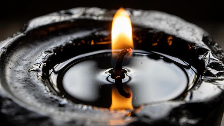 Burning charred wick curling and consuming oily fuel above shallow pool, reflecting orange flame