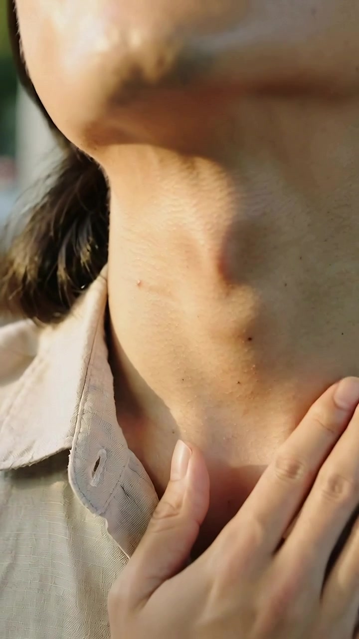 Video verticale: La mano destra scivola via dal riposo, donna che si controlla la gola all'aperto illuminato dal sole, con il colletto aperto.