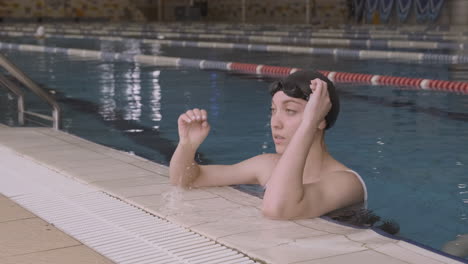 beautiful young girl leaning on the edge of the indoor pool puts on her swimming goggles