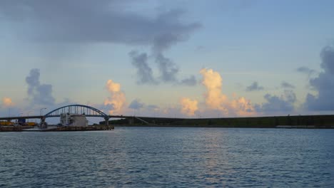 From-the-boat's-vantage-point,-a-charming-coastal-town-comes-into-view,-with-a-bridge-silhouetted-against-sunset-lit-clouds