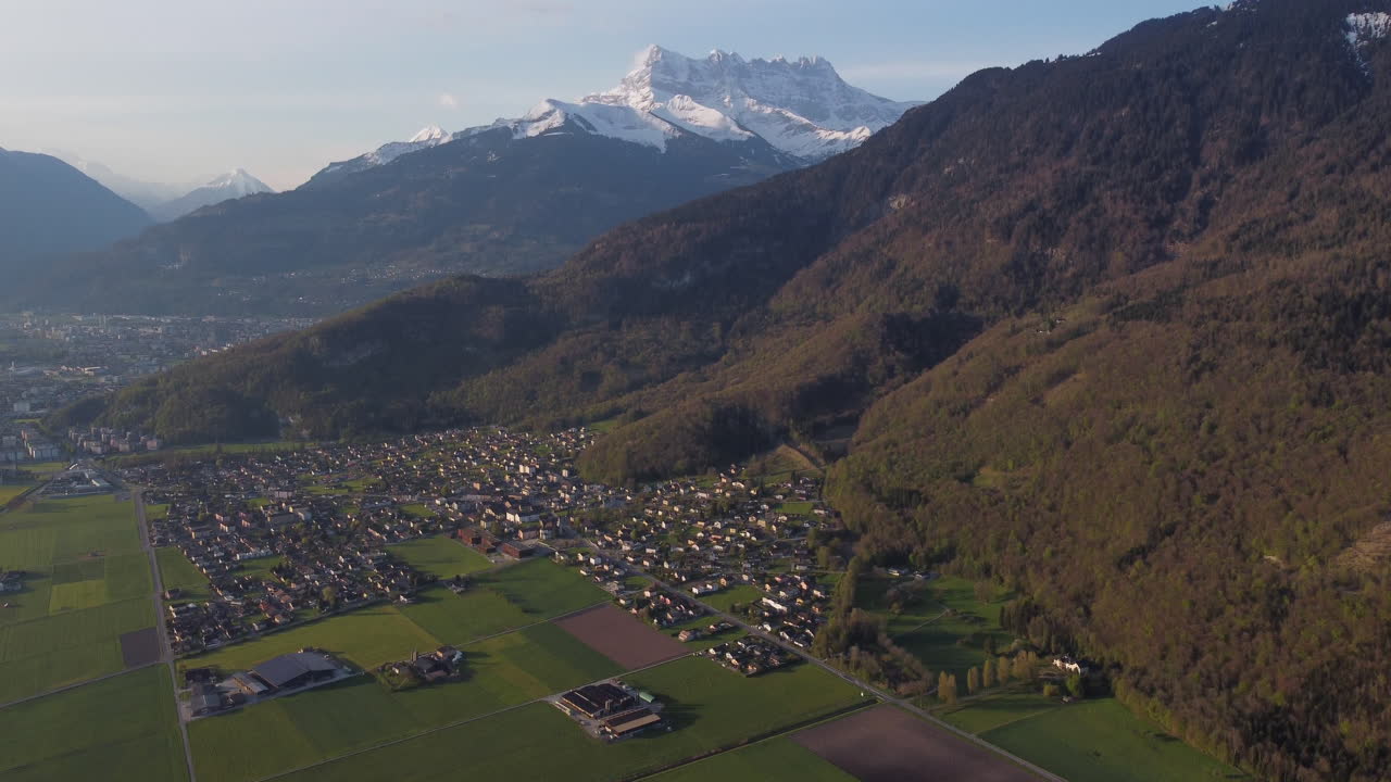 Aerial tilt up shot of farms and snow covered Alps in Vionnaz, Switzerland on a sunny day.