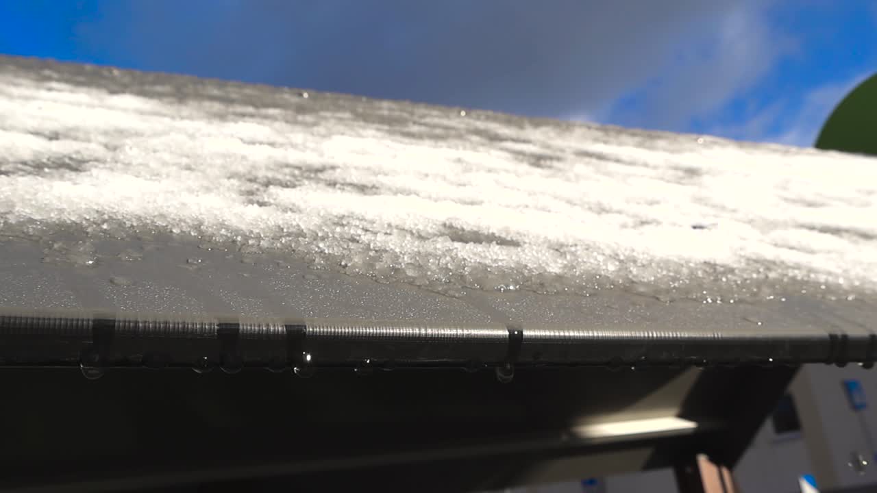 Bottom up close up view of a playground roof with white fluffy melting snow and ice on it during a sunny winter morning day. Water is flowing and dropping from the edge of the roof in slow motion