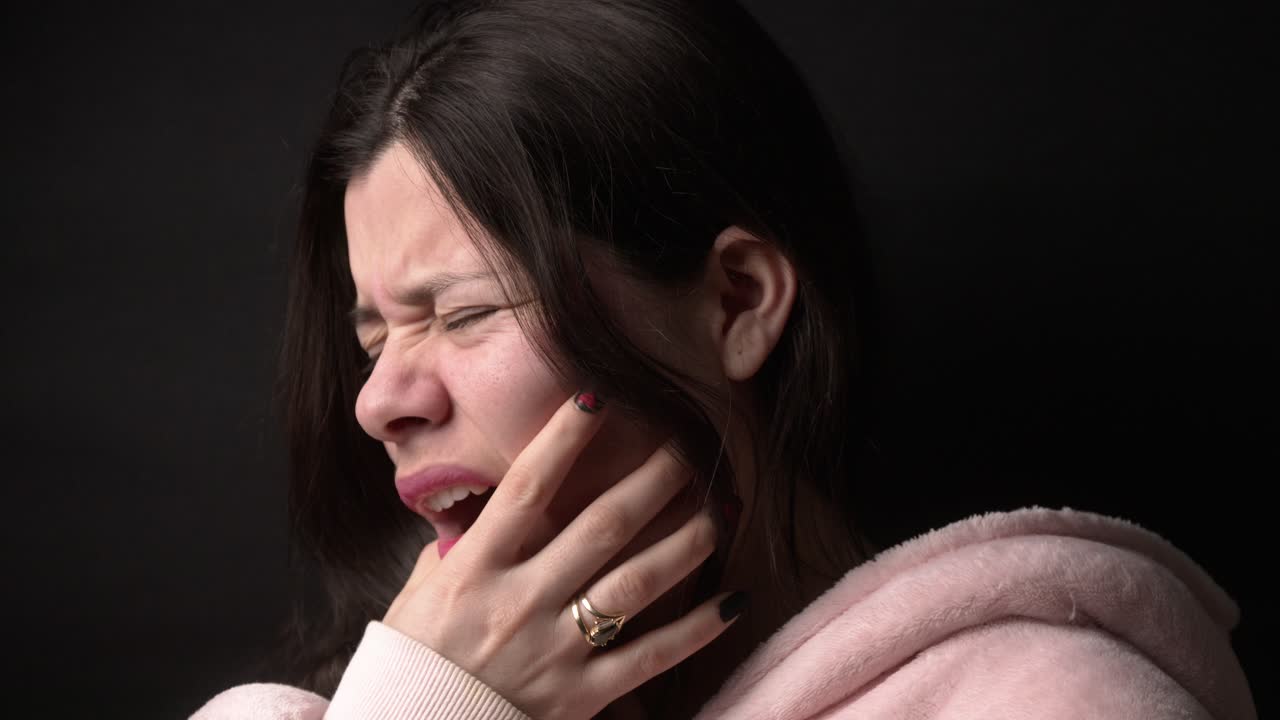 Close-up of a woman touching her cheek in discomfort, set against a dark background. The dramatic lighting highlights her expression, conveying intense emotion and vulnerability