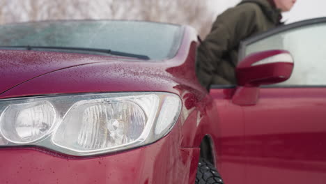 celebrity wearing winter jacket steps out of red car during snowy season with visible water droplets on vehicle surface and cold weather background