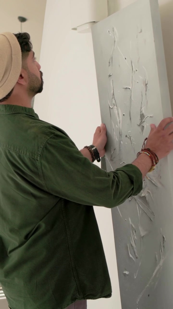 Video verticale: Uomo con berretto che prende una tela da un mobile TV basso a casa, appendendo un'opera d'arte per decorazione