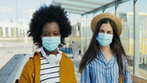 portrait of young african american and caucasian women travellers with backpack and wearing medical mask looking at camera outdoors