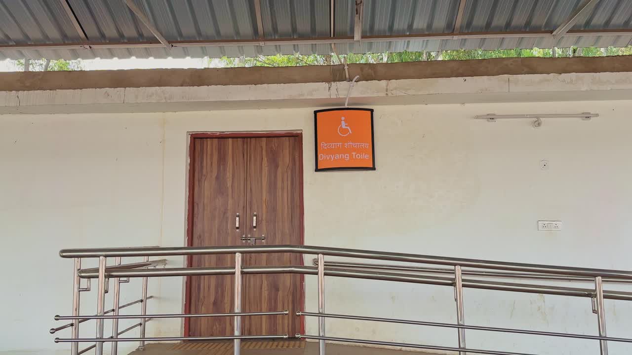 Plano de seguimiento lento a lo largo de una rampa que conduce a un baño accesible en la India, con pasamanos de acero inoxidable que guían el acceso en silla de ruedas hacia una entrada claramente marcada bajo una estructura cubierta
