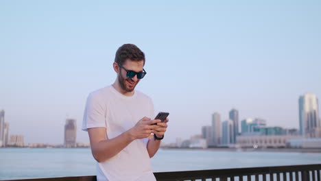 Un-Joven-Atractivo-Y-Apuesto-Con-Una-Hermosa-Sonrisa-Y-Una-Apariencia-Hipster-De-La-Generación-Milenaria-Z-Se-Desplaza-A-Través-De-Las-Redes-Sociales-Con-Risas-De-Teléfonos-Inteligentes-Y-Sonrisas-De-Noticias-Sobre-Aplicaciones.