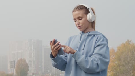 Joven-Deportista-Seria-Con-Capucha-Azul-Con-Auriculares-Mirando-El-Teléfono-Inteligente-Antes-De-Entrenar-En-El-Camino-Del-Bosque