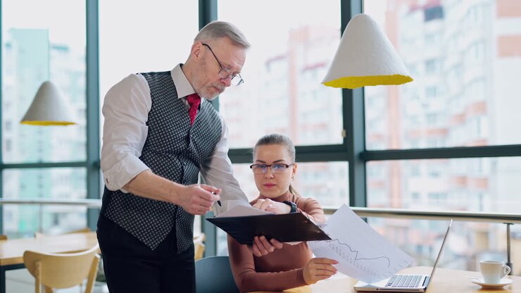 Moden forretningsmand forklarer arbejdet til en sekretær. Ung kvinde sidder ved sin arbejdsplads på kontoret og lytter til forretningsmanden.