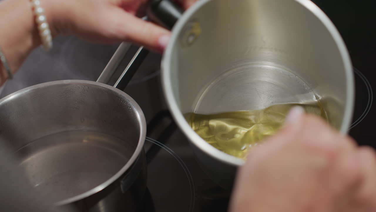 Close up of chef stirring thick brown-like melted substance inside aluminum pan using wooden stick beside pot of steaming water on stovetop, showcasing careful cooking technique