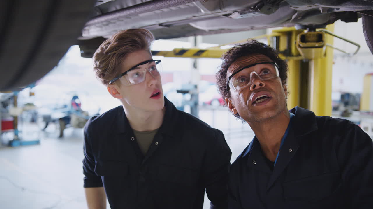Male Tutor With Student Looking Underneath Car On Hydraulic Ramp On Auto Mechanic Course