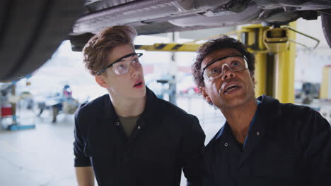 tutor masculino con estudiante mirando debajo del coche en la rampa hidráulica en el curso de mecánica de automóviles
