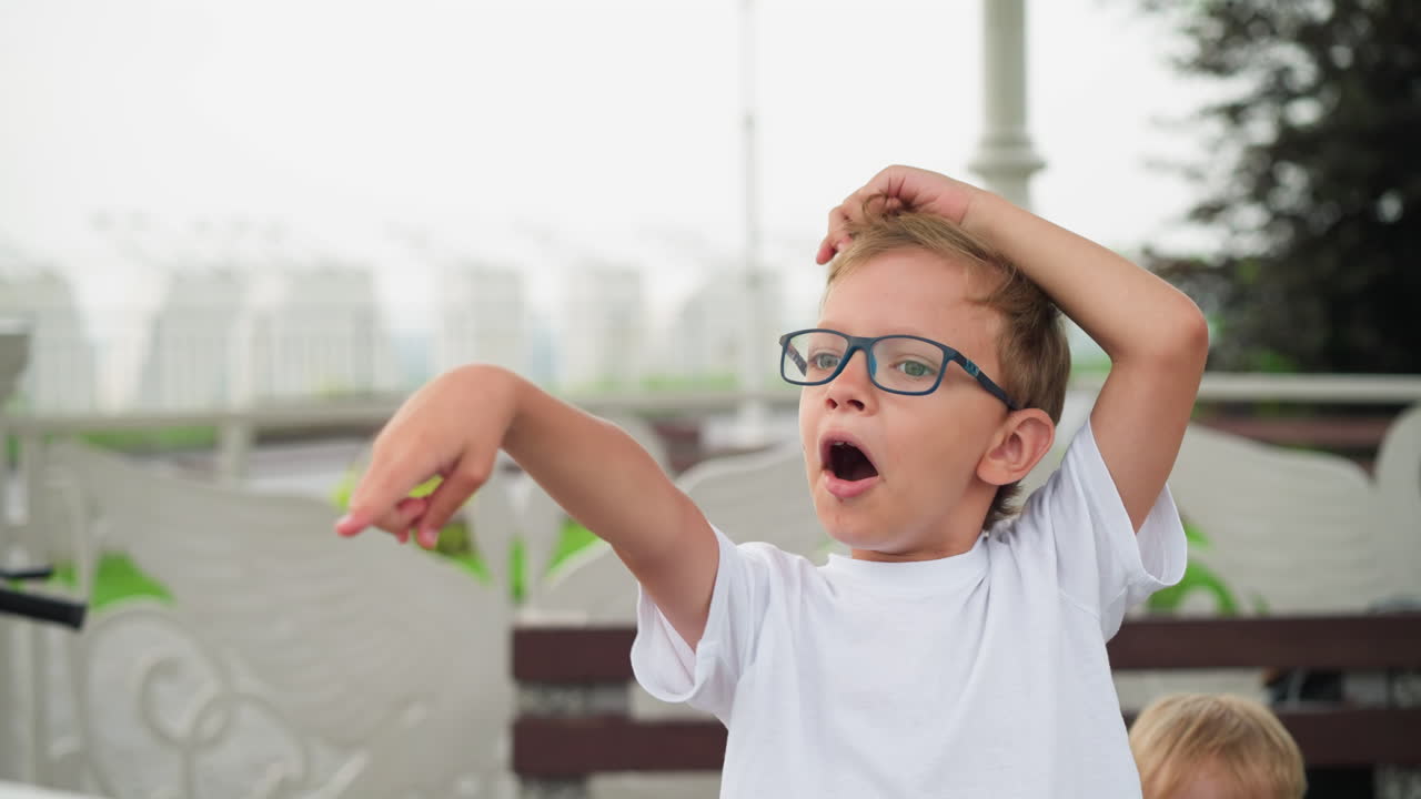A young boy wearing glasses playfully points at something in the distance while standing outdoors