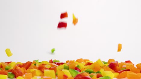 brightly colored red, green, yellow and orange diced bell pepper fresh vegetable pieces falling down onto white table top and bouncing into a pile in slow motion