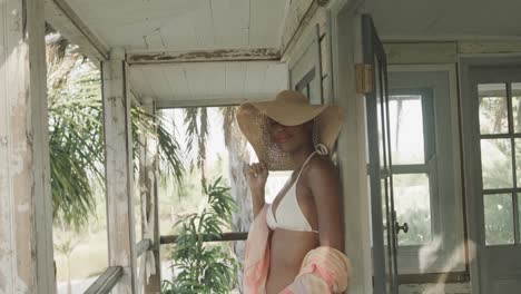 portrait of biracial woman in bikini and sunhat smiling on porch of wooden beach house, slow motion