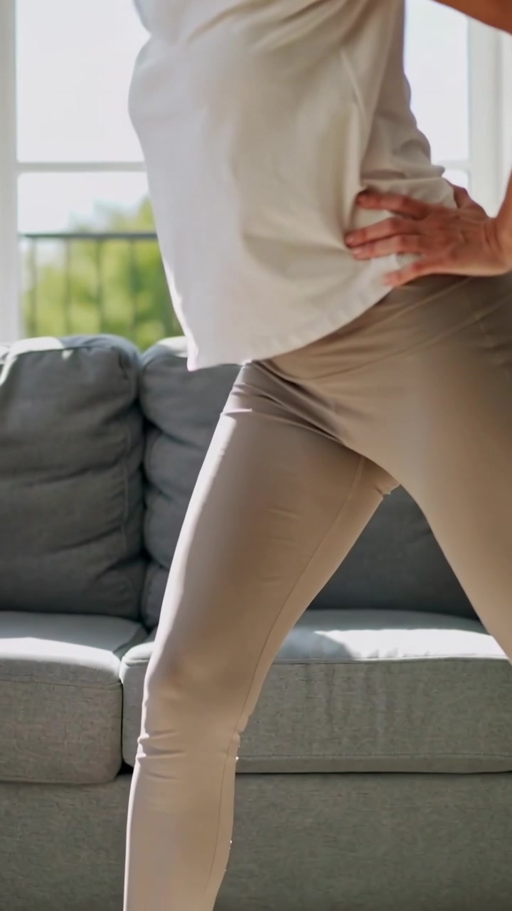 Vídeo vertical: Mulher começando a se alongar em casa com os pés afastados, de leggings, para flexibilidade, com sofá cinza.